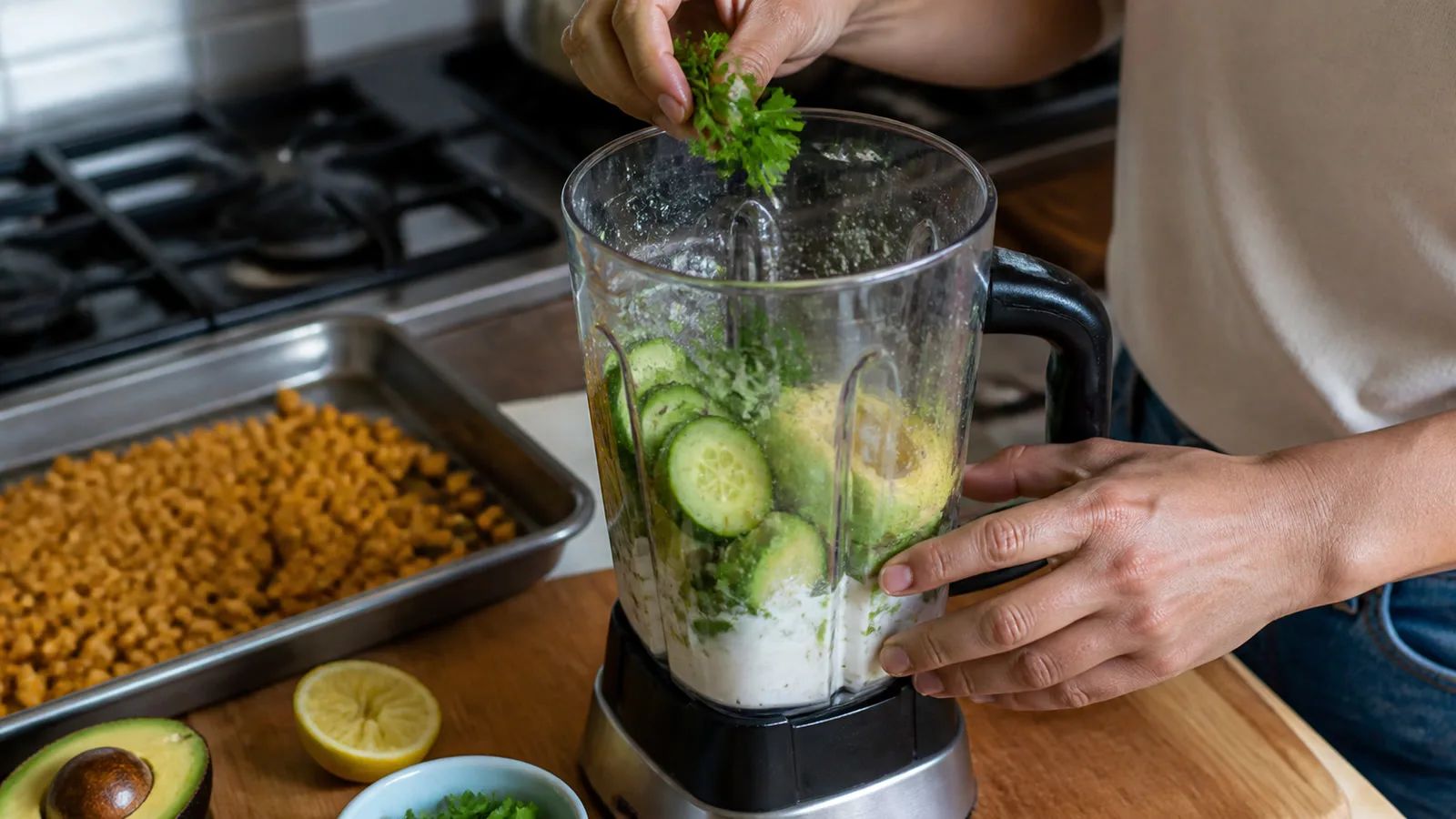 Manos agregando cilantro a una licuadora con pepino aguacate yogur y limon en cocina casera